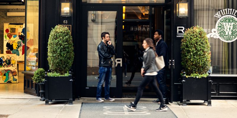 Two Men Stand in Front of the Entrance to Hollingsworth Luxury Apartments as a Woman Walks By