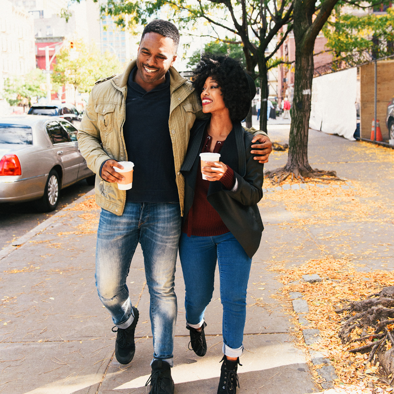A Young African-American Couple Smile As They Walk Together Down the Street With Hot Coffees
