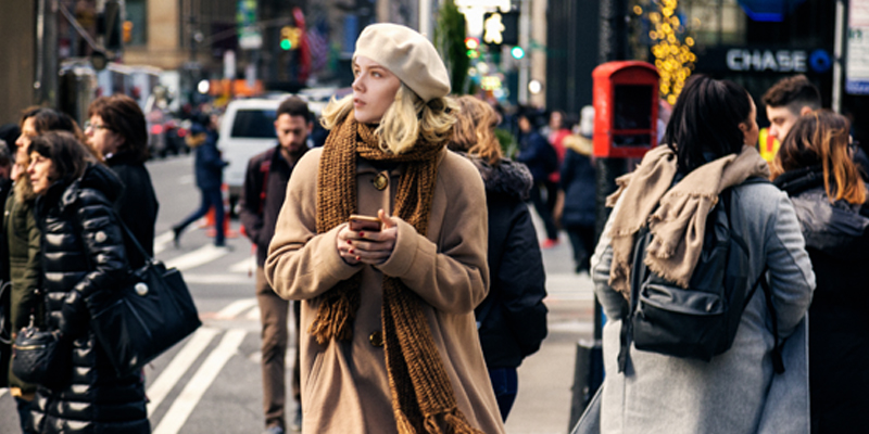 Young Girl With Short Blonde Hair and Brown Winter Gear Looks Up From Her Phone While Using a Busy City Crosswalk