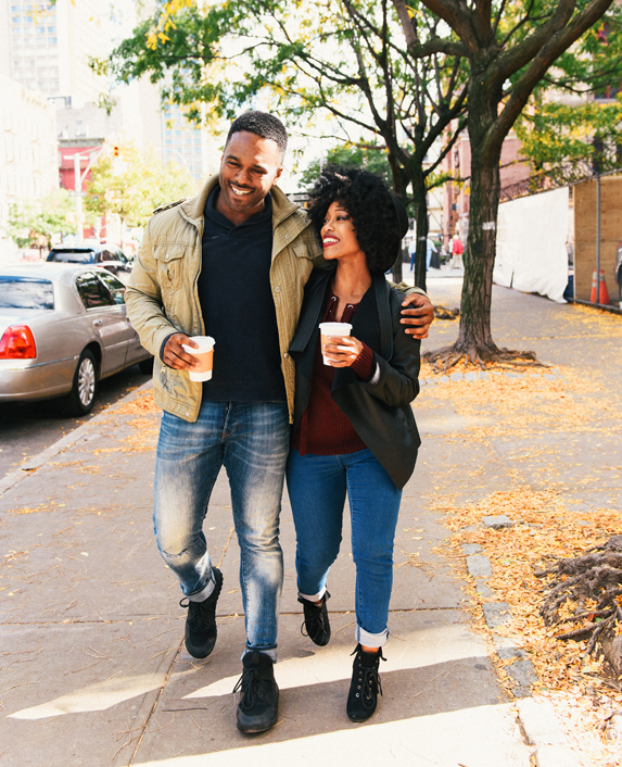 A Young African-American Couple Smile As They Walk Together Down the Street With Hot Coffees