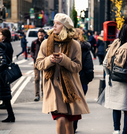 Young Girl With Short Blonde Hair and Brown Winter Gear Looks Up From Her Phone While Using a Busy City Crosswalk